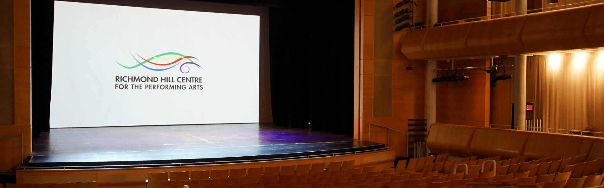 Image of an audience view of a theatre stage. On the stage, there is a projector screen showing the "Richmond Hill Centre for the Performing Arts" Logo.
